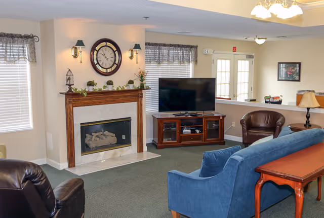 Cozy common room with a fireplace and clock, TV on a wooden cabinet, and seating including a blue sofa and leather chairs.