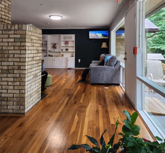 A senior woman sitting on a gray couch in a living room with wooden floors, a brick column, built-in white shelves, a floor lamp, and a wall-mounted TV. Large windows and a glass door provide a view of an outdoor patio area with greenery.