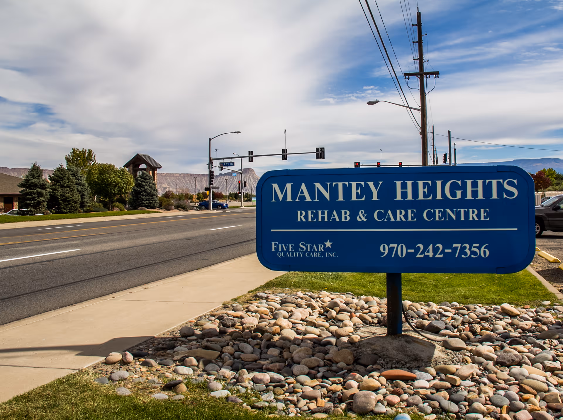 A blue sign for Mantey Heights Rehab & Care Centre located on a roadside with a sidewalk and rocks around its base. The sign includes the phone number 970-242-7356 and the text Five Star Quality Care, Inc. In the background, there are trees, a building, traffic lights, and a partly cloudy sky.