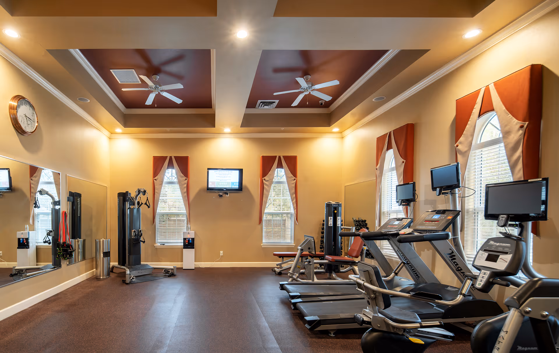 A well-lit fitness room with exercise equipment including treadmills, a recumbent bike, weight machines, and dumbbells. The room has beige walls, three tall windows with red and beige curtains, ceiling fans, and a wall-mounted TV. There is a large mirror on one wall and a clock above it.
