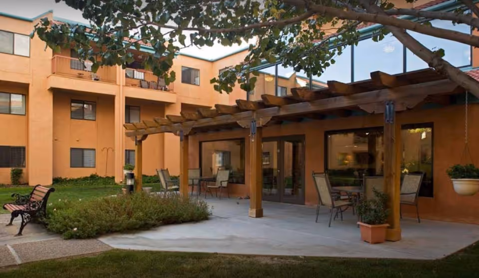 Outdoor patio area at Sandia Vista Senior Living with a wooden pergola, several chairs and tables, potted plants, a bench, and a tree with green leaves. The patio is adjacent to a multi-story building with large windows and a warm orange exterior.