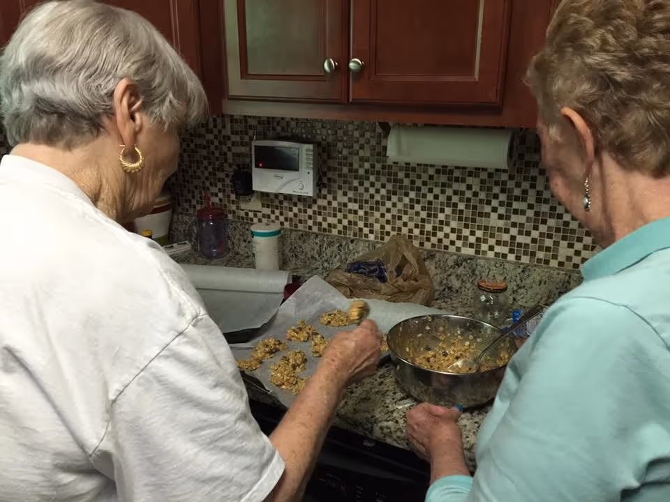 Two elderly women are in a kitchen preparing cookies. One woman is placing cookie dough onto a baking sheet lined with parchment paper, while the other is holding a mixing bowl with cookie dough. The kitchen has a granite countertop, mosaic tile backsplash, and wooden cabinets.