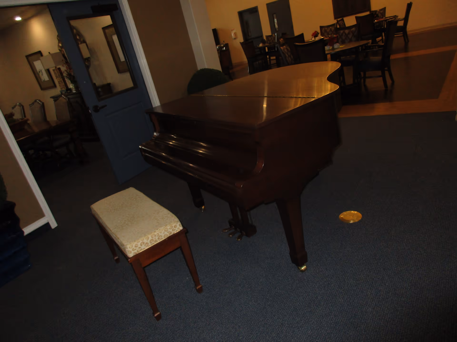 A dark wood grand piano with a matching cushioned bench sits on a blue carpeted floor in a room. In the background, there is a blue door leading to another room with chairs and tables visible. The room has a warm, cozy ambiance with some framed pictures on the walls.