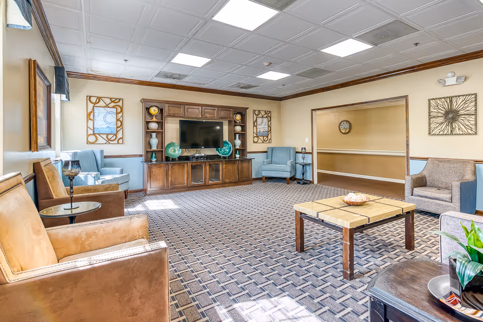 A spacious senior living common room with armchairs, a TV cabinet, coffee table, and patterned carpet.