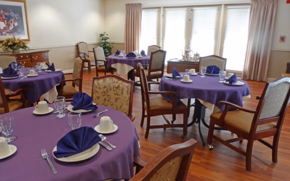 Dining room with round tables covered in purple tablecloths and white underlays, each set with plates, cups, glasses, silverware, and folded purple napkins. The room has wooden floors, upholstered chairs, large windows with beige curtains, and a sideboard with a floral arrangement.