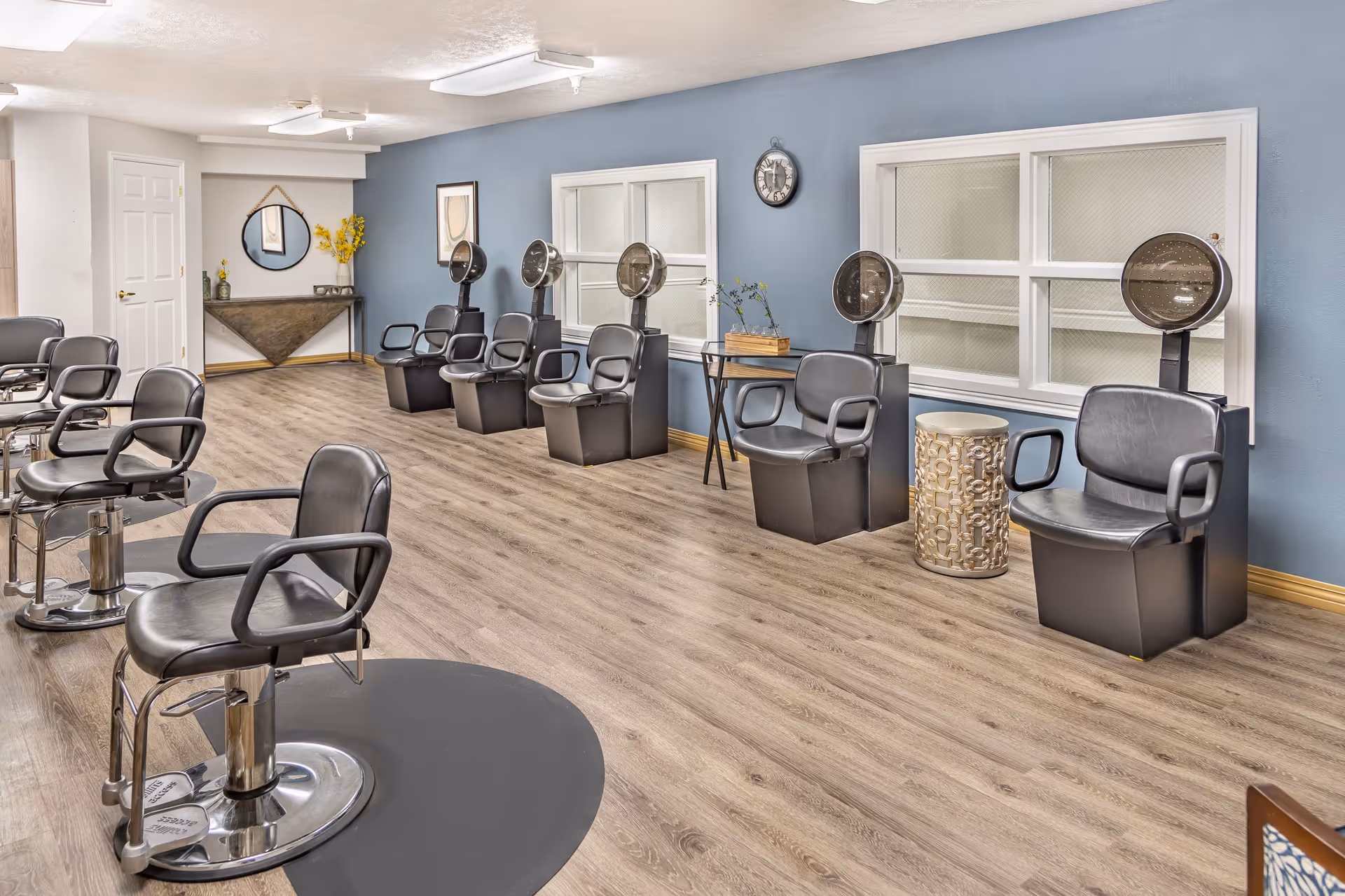 Salon-style common room with rows of black styling chairs and hooded hair dryers against a blue wall on wood-look flooring.