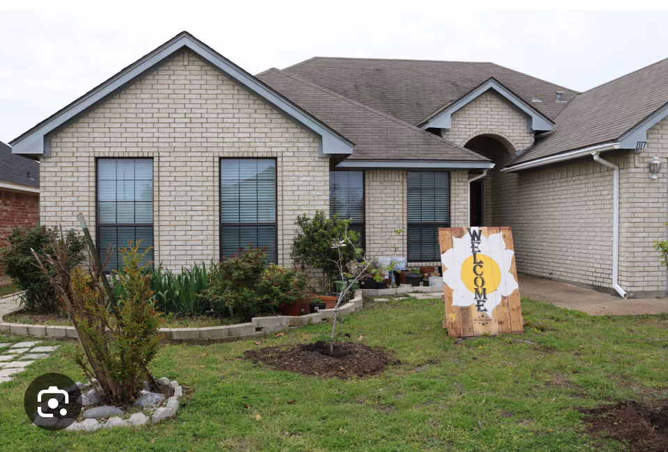Front exterior of a single-story brick house with a small lawn, garden beds, and a wooden 'WELCOME' sign propped in the yard.