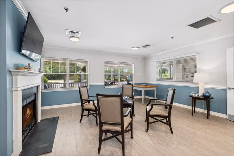 A bright and clean common area with four chairs arranged around a small table, a fireplace with a mounted TV above it, large windows letting in natural light, and a side table with a lamp and decorative items.