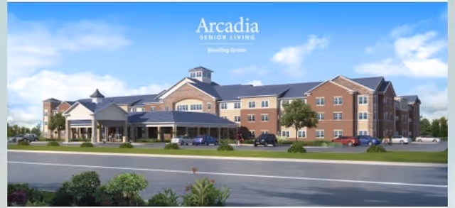 Exterior view of a large, multi-story senior living facility building with a brick and beige facade, multiple windows, a covered entrance, and a parking area with several cars. The sky is clear with some clouds.