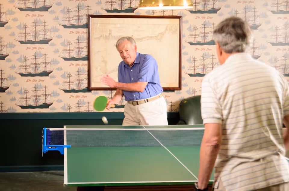 Two elderly men playing table tennis indoors. One man in a blue shirt is hitting the ball with a green paddle, while the other man in a striped shirt is facing him. The room has wallpaper with ship illustrations and a framed map on the wall.