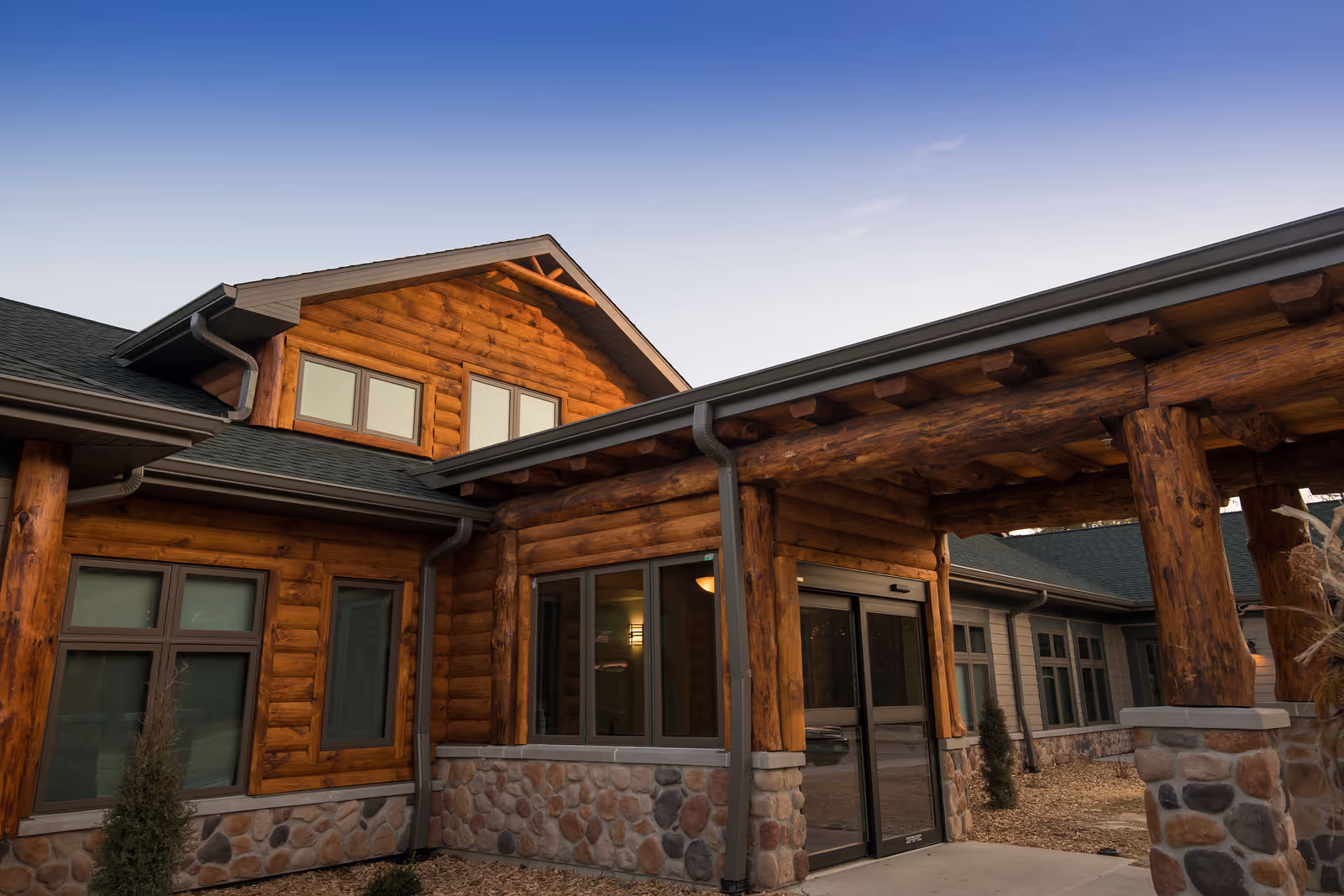 Exterior front entrance of a log-style senior living building with stone foundation and heavy wooden columns under a covered walkway.
