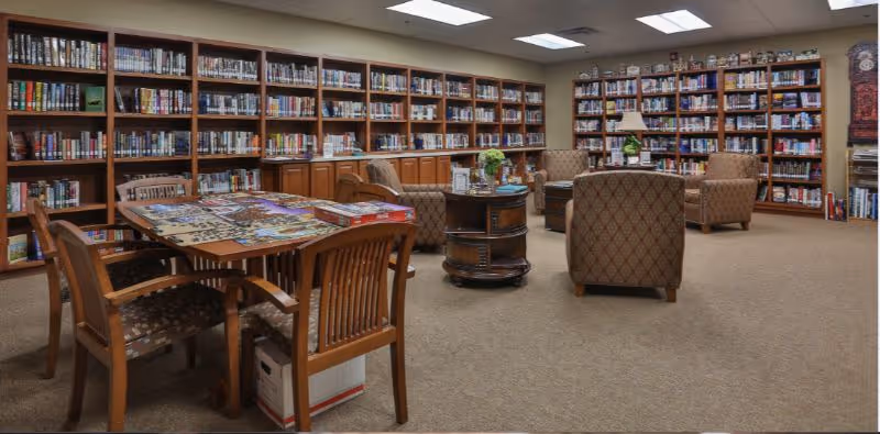 Spacious interior library with floor-to-ceiling bookshelves, a table with a jigsaw puzzle and chairs, and upholstered seating around small tables.