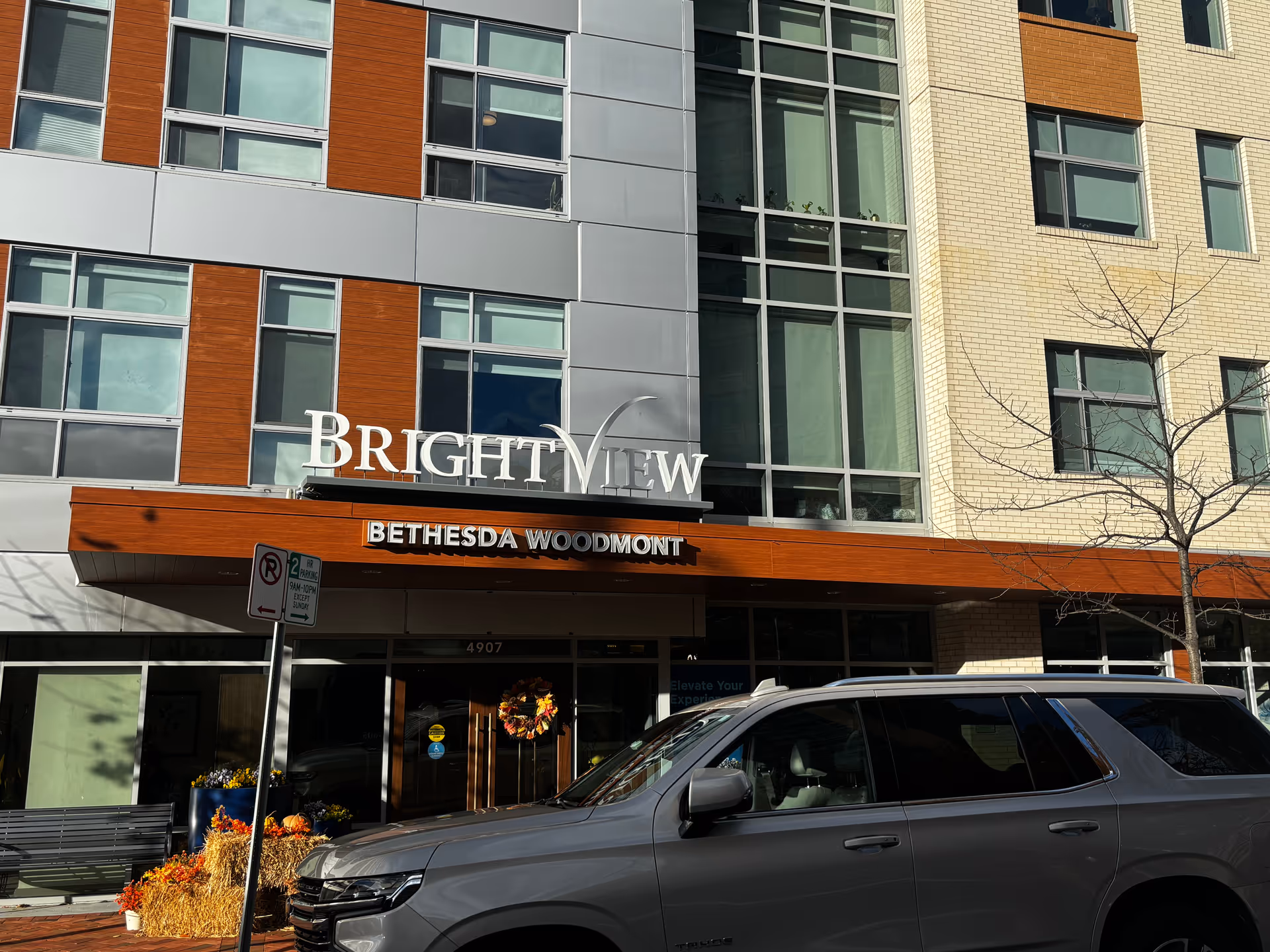 Exterior view of the Brightview Bethesda Woodmont building with a modern facade featuring large windows, a mix of gray, brown, and beige panels, and a sign displaying the facility name. A silver SUV is parked in front, and there is a small tree without leaves on the right side.