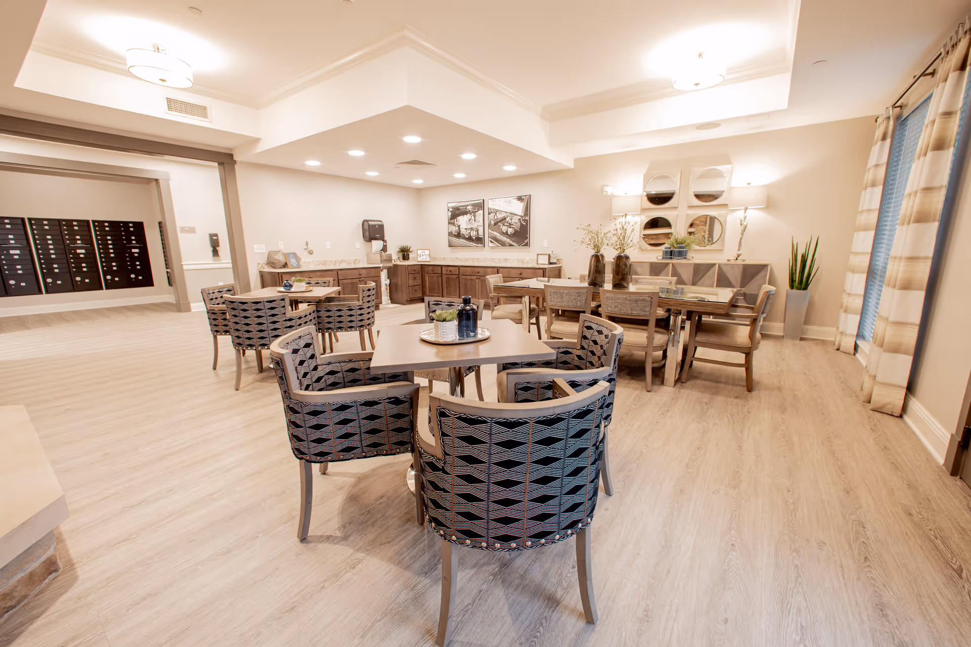 A bright and spacious dining area in a senior living facility with multiple tables and cushioned chairs arranged neatly. The room features light wood flooring, soft beige walls, and large windows with striped curtains allowing natural light. Decorative elements include vases with plants, wall art, and mirrors. In the background, there is a counter area with cabinets and a paper towel dispenser.