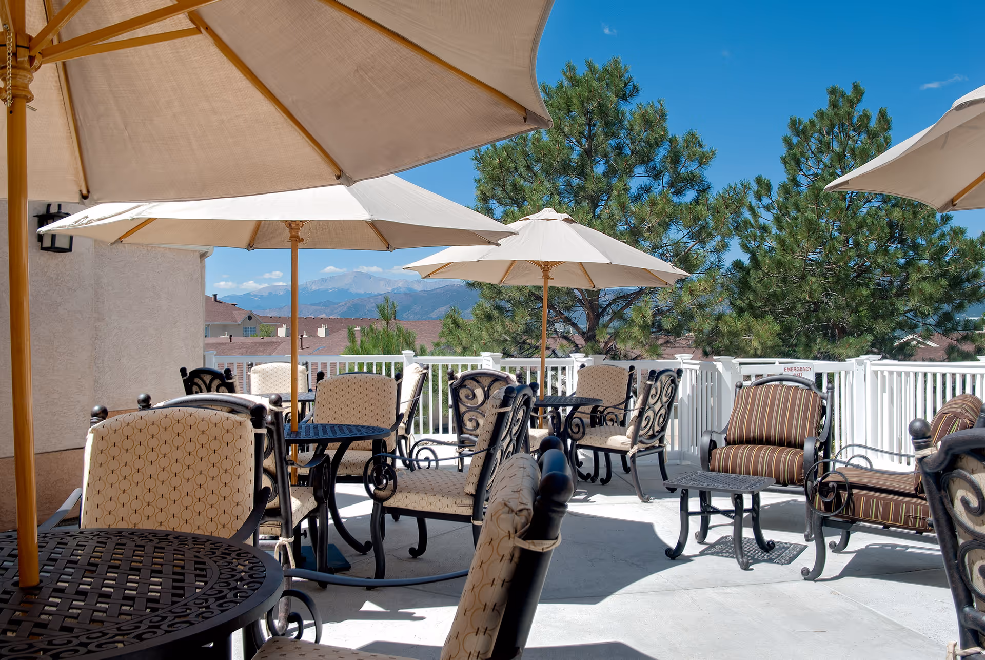 Outdoor patio area with multiple tables and cushioned chairs under large beige umbrellas. The patio is surrounded by a white railing, with trees and mountains visible in the background under a clear blue sky.