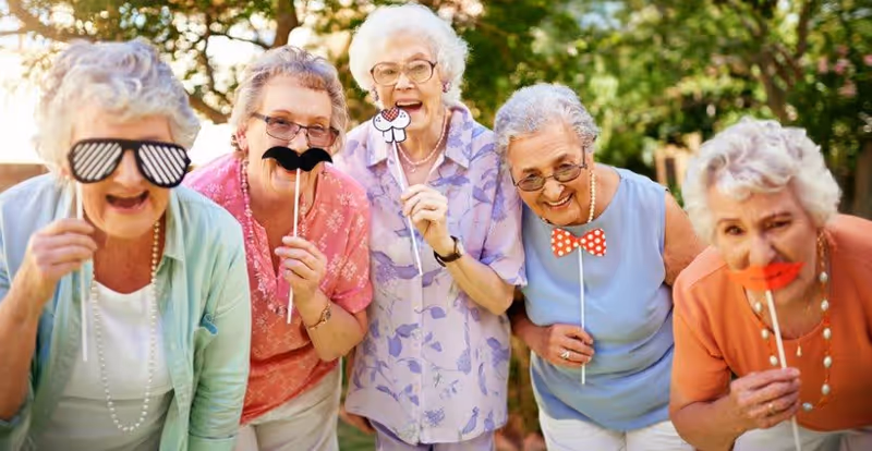 Five elderly women standing outdoors in a garden, each holding playful photo props such as sunglasses, mustaches, a cupcake, a bow tie, and red lips, smiling and enjoying a sunny day.