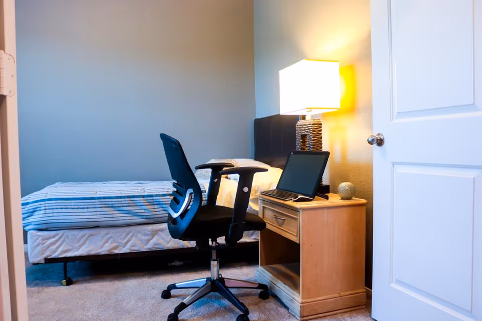 A furnished bedroom with a bed, desk, chair, laptop, and a lamp.
