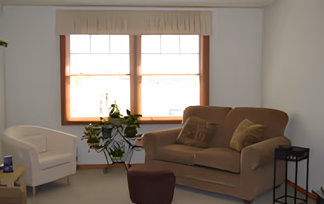 A cozy living room with a beige loveseat adorned with two pillows, a white armchair, a small brown ottoman, and a decorative metal plant stand with green plants in front of two large windows with beige valances.
