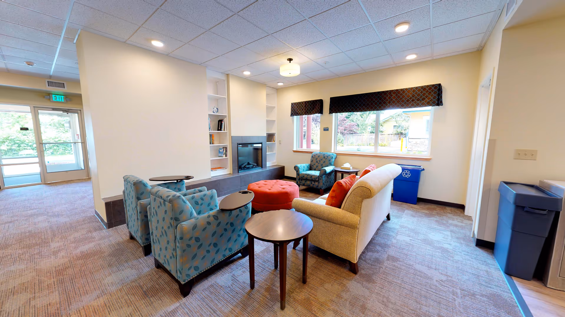 A cozy seating area in an assisted living facility with two blue patterned armchairs, a beige sofa with red pillows, a round red ottoman, and small wooden tables. The room has large windows with dark valances, a built-in fireplace with shelves, and a carpeted floor. There is a recycling bin near the window and an exit door visible in the background.