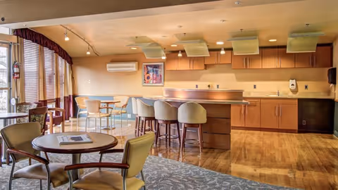 A spacious, well-lit common area with a kitchen counter and bar stools, several tables and chairs, large windows with curtains, and wooden flooring. The room has a warm color palette and modern lighting fixtures.