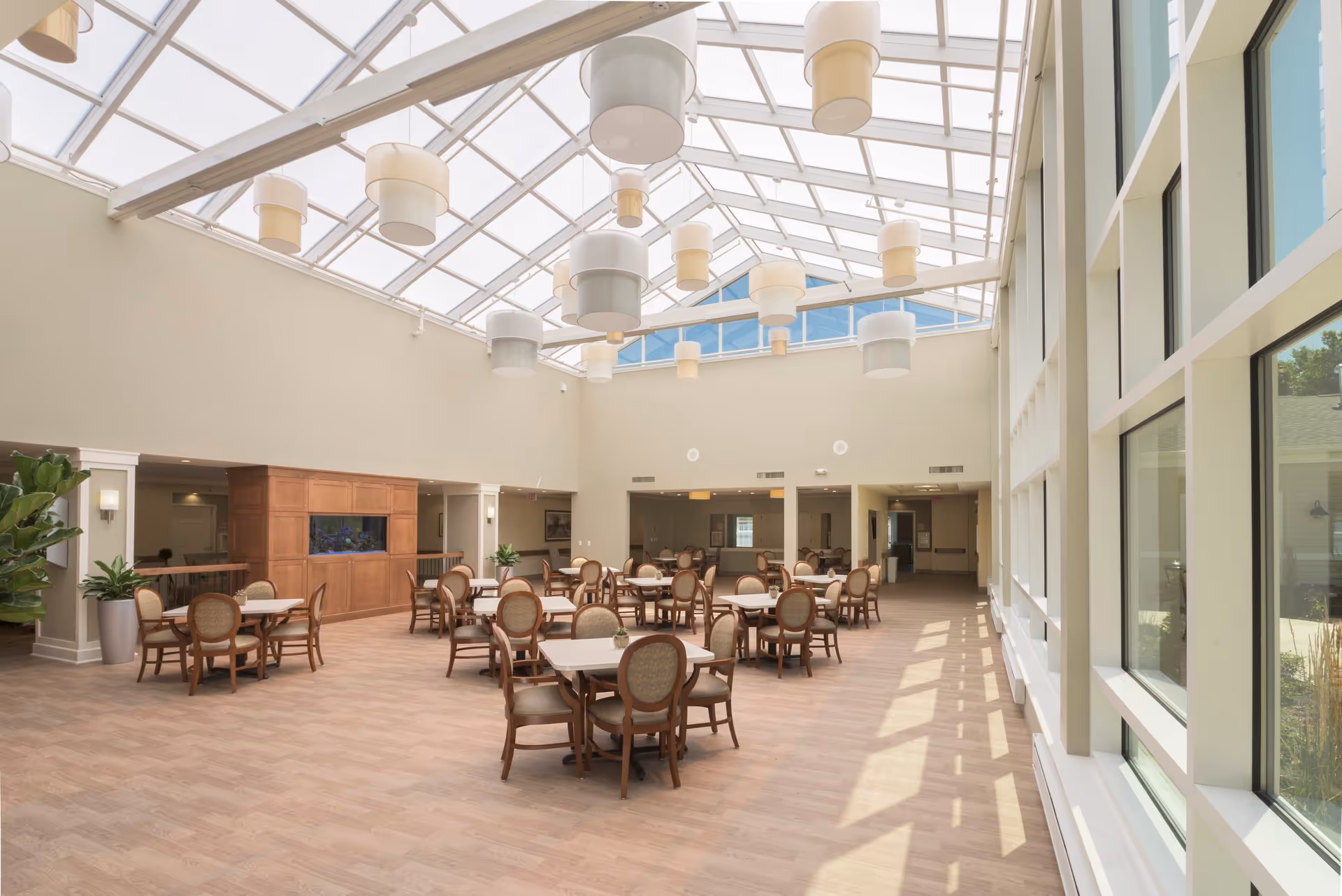 A spacious dining room with multiple tables and chairs arranged neatly. The room features a high glass ceiling with modern hanging light fixtures and large windows on one side allowing natural light to fill the space. There are plants and a wooden cabinet with an aquarium built in along one wall.