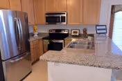 Modern kitchen with granite countertops, stainless steel refrigerator, oven, microwave, wooden cabinets, double sink, and a window with blinds.