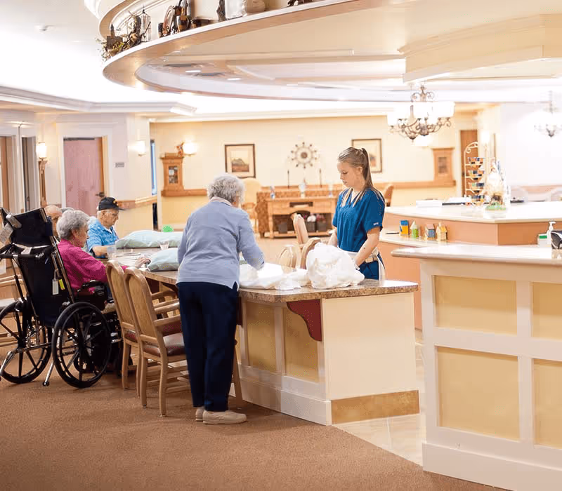 A senior living facility common area with elderly residents sitting around a table, one in a wheelchair, and a caregiver standing nearby assisting with folding linens. The room is warmly lit with beige walls, wooden furniture, and decorative items on shelves and walls.