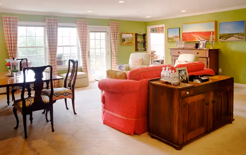 A bright and cozy living room with green walls and beige carpet. The room features a red upholstered sofa, a wooden sideboard with decorative items, a dining table with four chairs, and large windows with plaid curtains letting in natural light. Several framed landscape paintings hang on the walls.
