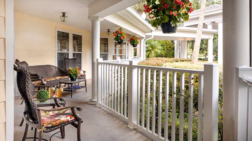Covered porch with wicker chairs and bench, floral cushions, hanging flower baskets, and a white railing overlooking shrubs.