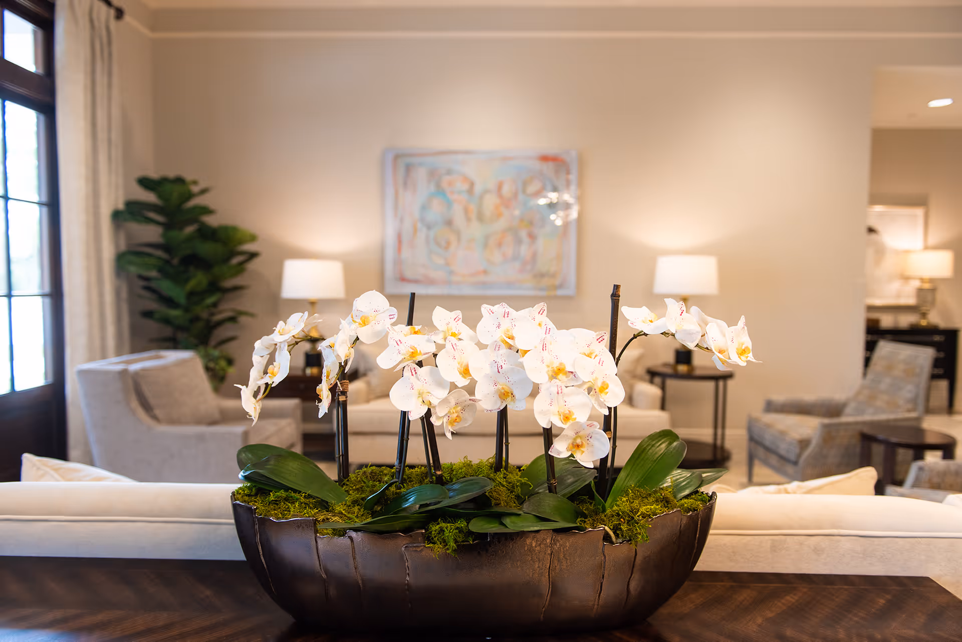 White orchid arrangement in a decorative planter on a table in a bright living room with sofas, chairs, lamps, and artwork.