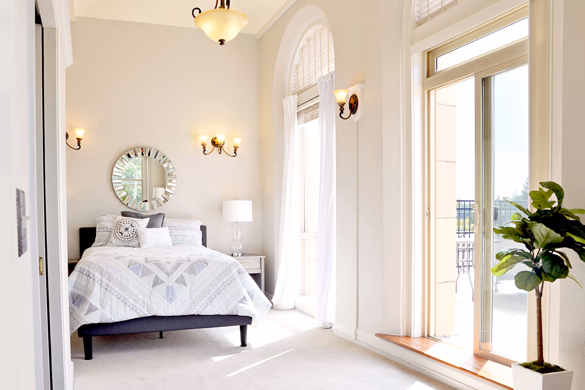 Bright bedroom with a neatly made bed featuring patterned bedding, a round decorative mirror on the wall above the bed, wall-mounted light fixtures, a bedside table with a lamp, large windows with white curtains, a sliding glass door leading to an outdoor patio, and a potted plant near the door.