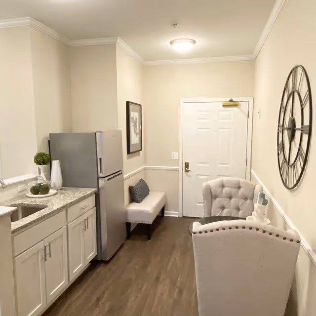 A small interior space featuring a kitchenette with white cabinets and a granite countertop, a stainless steel refrigerator, a small bench with a cushion, two upholstered chairs, a large decorative wall clock, and a white door at the end of the room.