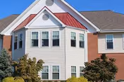 Exterior view of a residential building with white siding, red roof accents, multiple windows, and surrounding greenery under a clear blue sky.