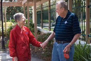 An elderly woman in a red jacket and an elderly man in a navy striped polo shirt holding hands and smiling at each other while standing outside near a garden and a fence.