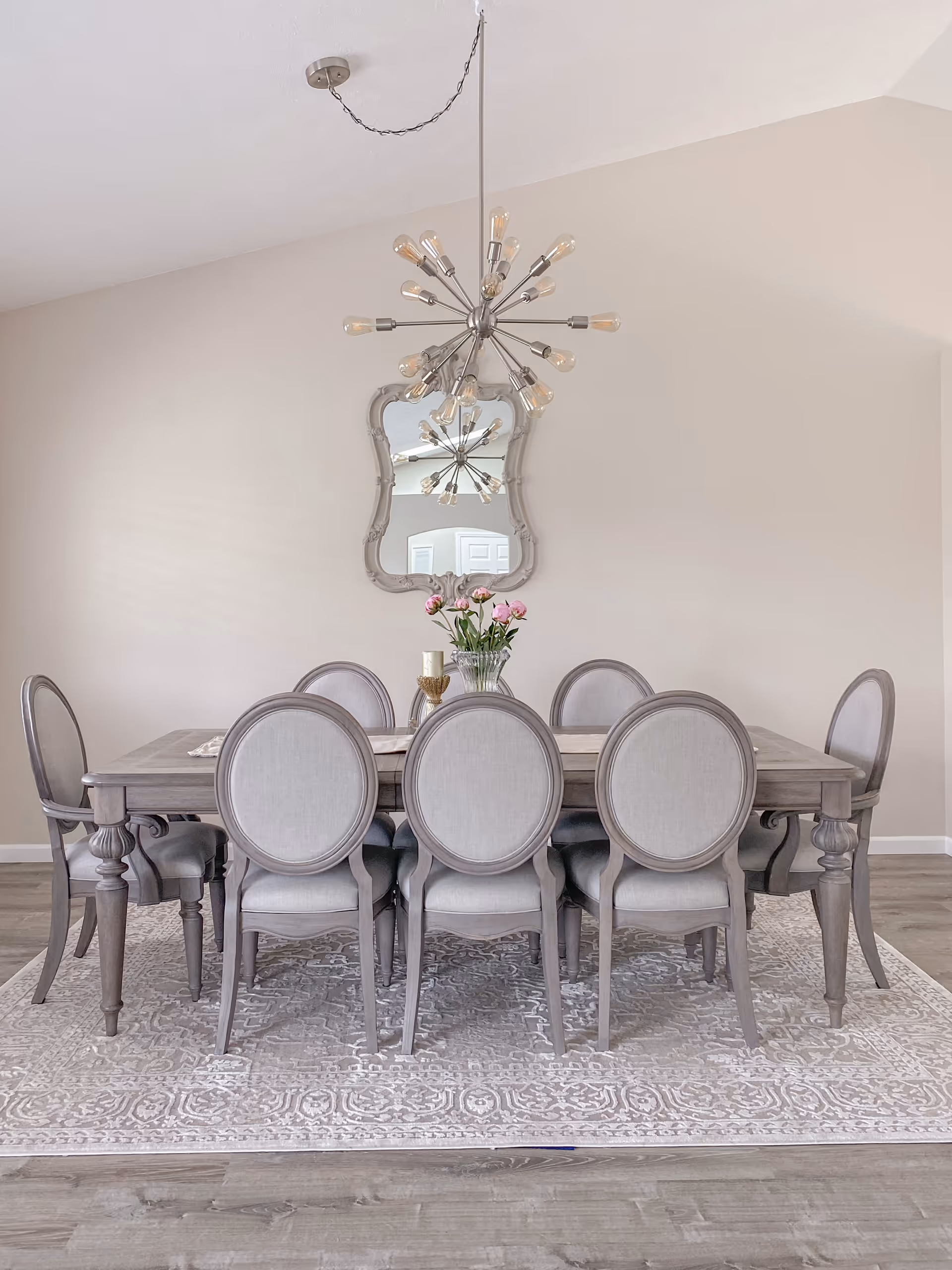 A dining room with a rectangular wooden table surrounded by eight upholstered chairs with rounded backs. A decorative mirror hangs on the beige wall behind the table, reflecting a modern chandelier with multiple exposed bulbs. A vase with pink flowers and a candle holder are placed on the table, which sits on a patterned area rug over wooden flooring.