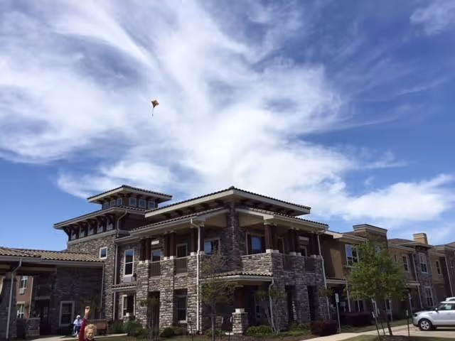 Exterior view of a multi-story senior living facility building with stone and brick facade under a partly cloudy blue sky. A person is flying a kite near the building, and there are some trees and parked cars visible.