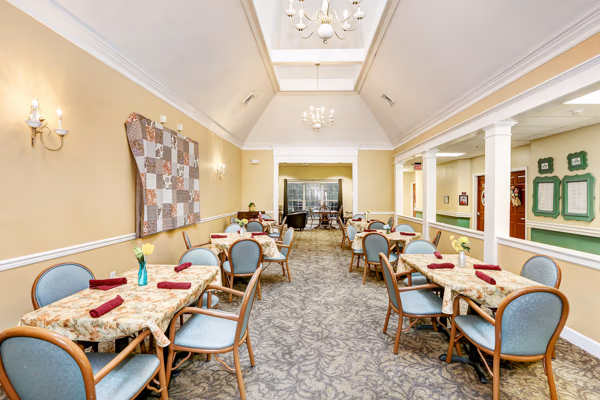 A bright dining room with several tables covered in floral tablecloths and set with red napkins and small flower vases. The room has beige walls, a patterned carpet, and two chandeliers hanging from a vaulted ceiling. There is a quilt hanging on one wall and framed notices on another. The room extends into a sitting area with large windows at the far end.