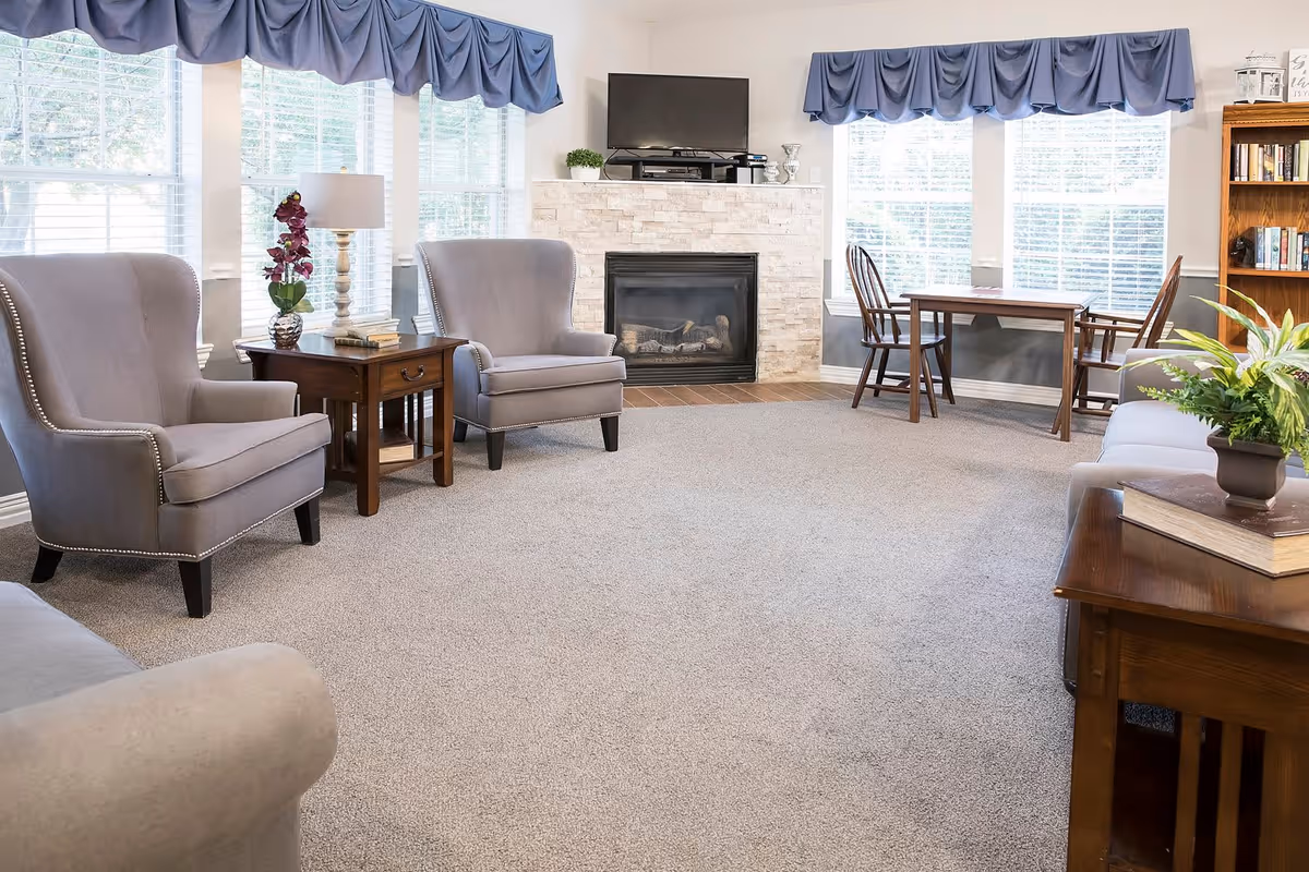 A bright living room with beige carpet, two gray upholstered armchairs, a wooden side table with a lamp and a purple orchid, a stone fireplace with a TV mounted above it, a wooden table with two chairs near large windows with blue valances, and a wooden bookshelf filled with books.