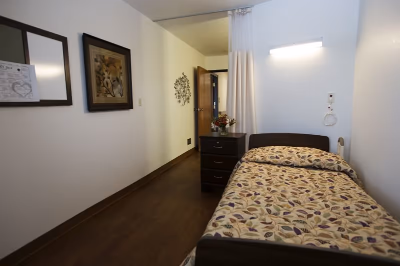 A small bedroom in a senior living facility with a single bed covered in a floral patterned bedspread. Next to the bed is a dark wooden nightstand with three drawers and a vase of flowers on top. The room has light-colored walls, a wall-mounted light above the bed, and a curtain that can be drawn for privacy. There are two framed pictures on the wall and an open door leading to a hallway.