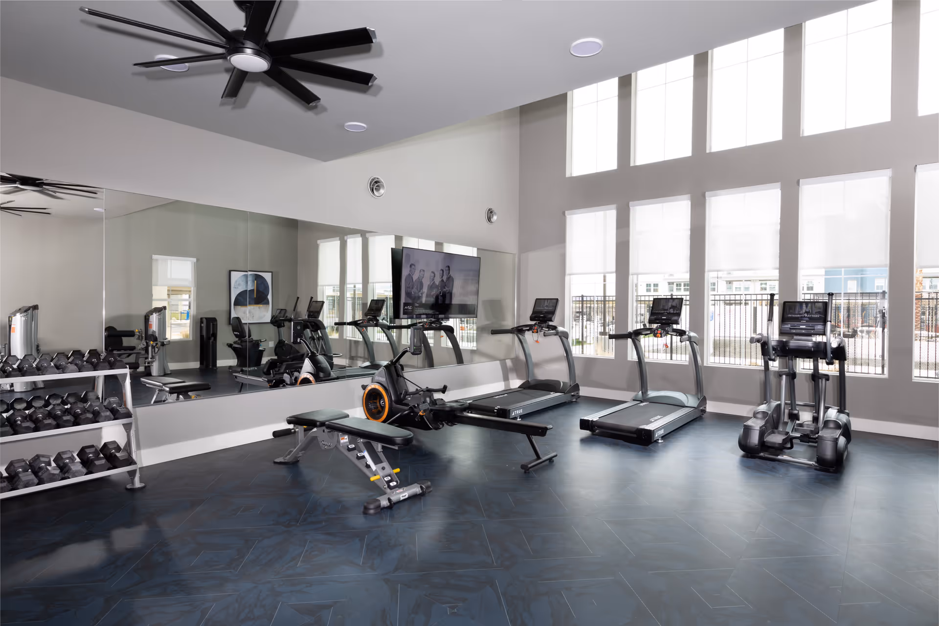 A bright and modern fitness room with large windows allowing natural light to fill the space. The room features two treadmills, an elliptical machine, a rowing machine, a weight bench, and a rack of dumbbells. A large mirror covers one wall, reflecting the equipment and enhancing the spacious feel. Ceiling fans are mounted on the ceiling for ventilation.