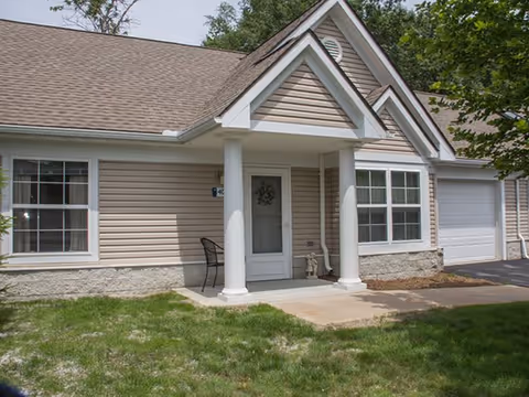 Exterior view of a single-story residential building with beige siding, white trim, and a gabled roof. The entrance features a small covered porch supported by two white columns, a white door with a decorative wreath, and a black metal chair to the left. There is a window on each side of the door and a garage door to the right. The lawn in front is green with some patches of dirt, and trees are visible in the background.