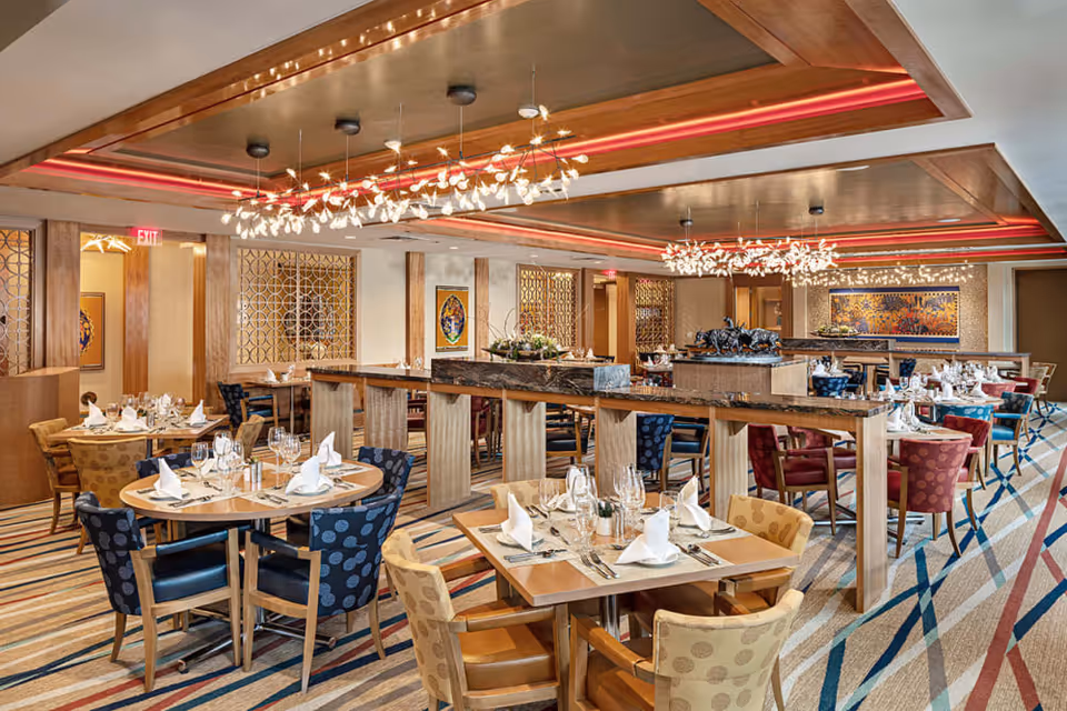 A spacious and elegantly decorated dining room with multiple tables set with white napkins, glassware, and silverware. The room features colorful patterned chairs, modern chandeliers with leaf-like designs, wooden accents, and a striped carpet. Artwork and decorative partitions adorn the walls.