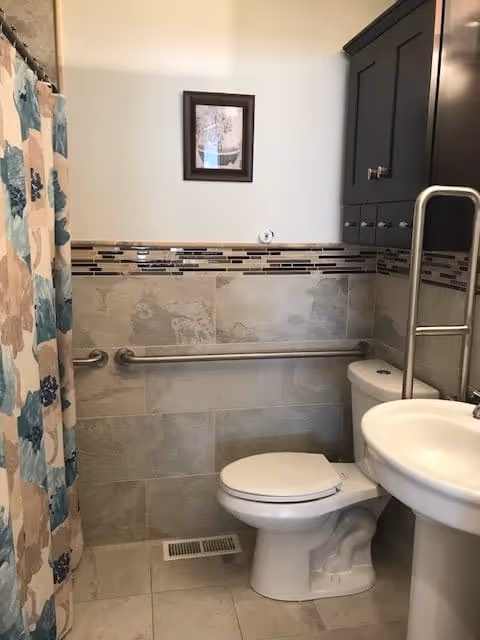 A small bathroom featuring a white toilet and pedestal sink. There is a grab bar installed horizontally along the tiled wall behind the toilet. The wall has beige tiles with a decorative horizontal strip of smaller tiles in various shades of brown and black. A shower curtain with a floral pattern in blue, beige, and white colors is partially visible on the left side. Above the toilet, there is a dark-colored cabinet with multiple drawers and a framed picture hanging on the wall.