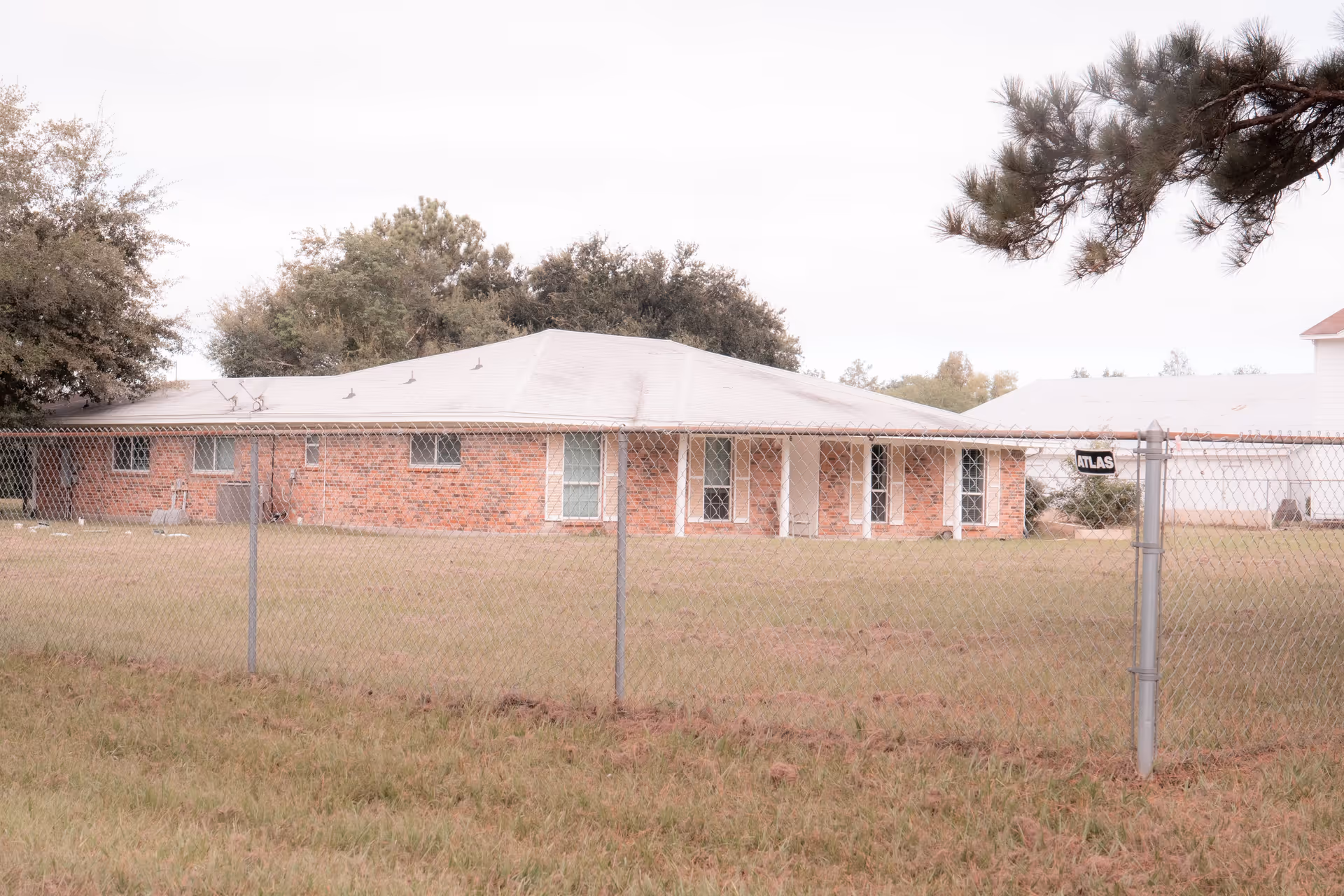 Single-story brick building viewed from outside behind a chain-link fence with a grassy yard and trees.