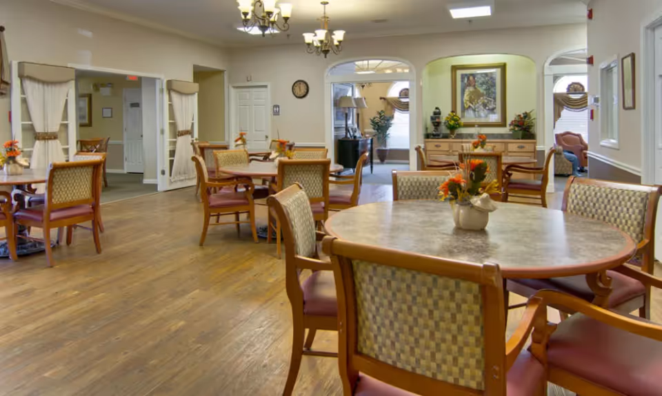 A spacious dining room in a senior living facility with multiple round tables and cushioned chairs arranged neatly. Each table has a small floral centerpiece. The room features wooden flooring, soft lighting from chandeliers, and a sideboard with decorative items and a framed painting on the wall. There are archways leading to other rooms and windows with blinds and valances.