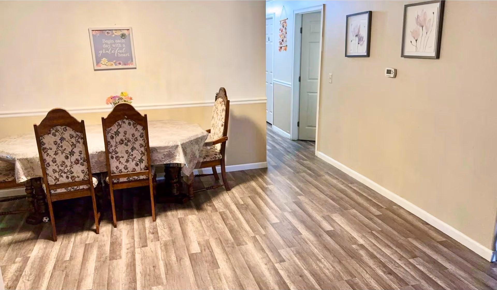 A dining area with a wooden table covered by a floral tablecloth and four wooden chairs with floral upholstery. On the wall above the table is a framed sign that reads 'Begin each day with a grateful heart.' The floor is wood-patterned, and the walls are painted beige with white trim. There is a hallway visible in the background with two framed floral pictures on the right wall.