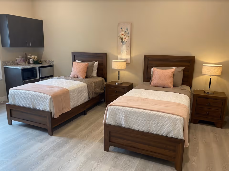 Two twin beds with wooden frames and pink-accented bedding in a furnished bedroom with nightstands, lamps, and a small kitchenette area.