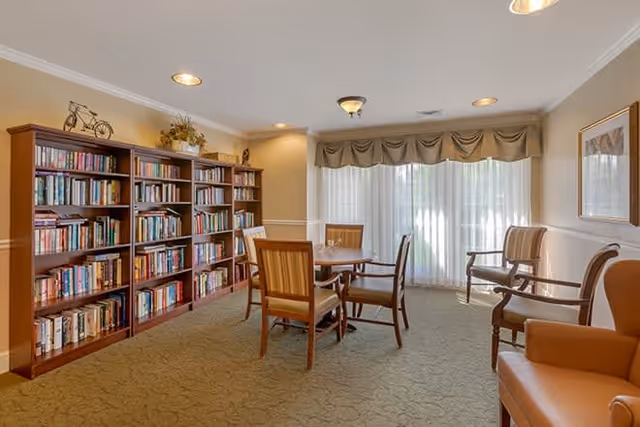 A cozy reading room with a large wooden bookshelf filled with books along one wall, a round wooden table surrounded by four chairs in the center, and additional seating including armchairs near a window with sheer curtains and a valance, illuminated by ceiling lights.