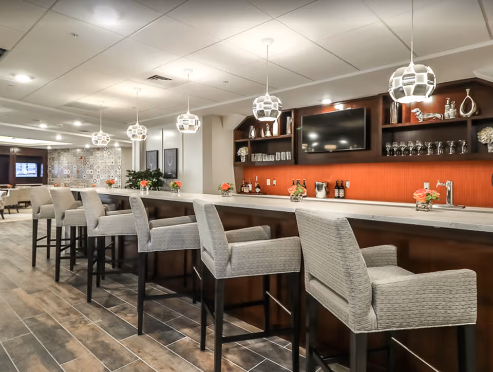 Modern communal bar and lounge with a long marble-top counter, upholstered high stools, pendant lights, and a wall-mounted TV.