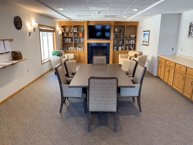A senior living common room with a central table surrounded by chairs, bookshelves, a TV above a fireplace, and seating along the walls.
